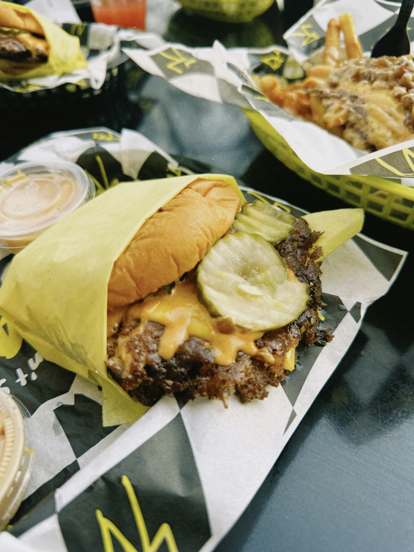 A smash burger with melted cheese and pickles, the patty edge crisped brown, wrapped in paper next to a cup of sauce.