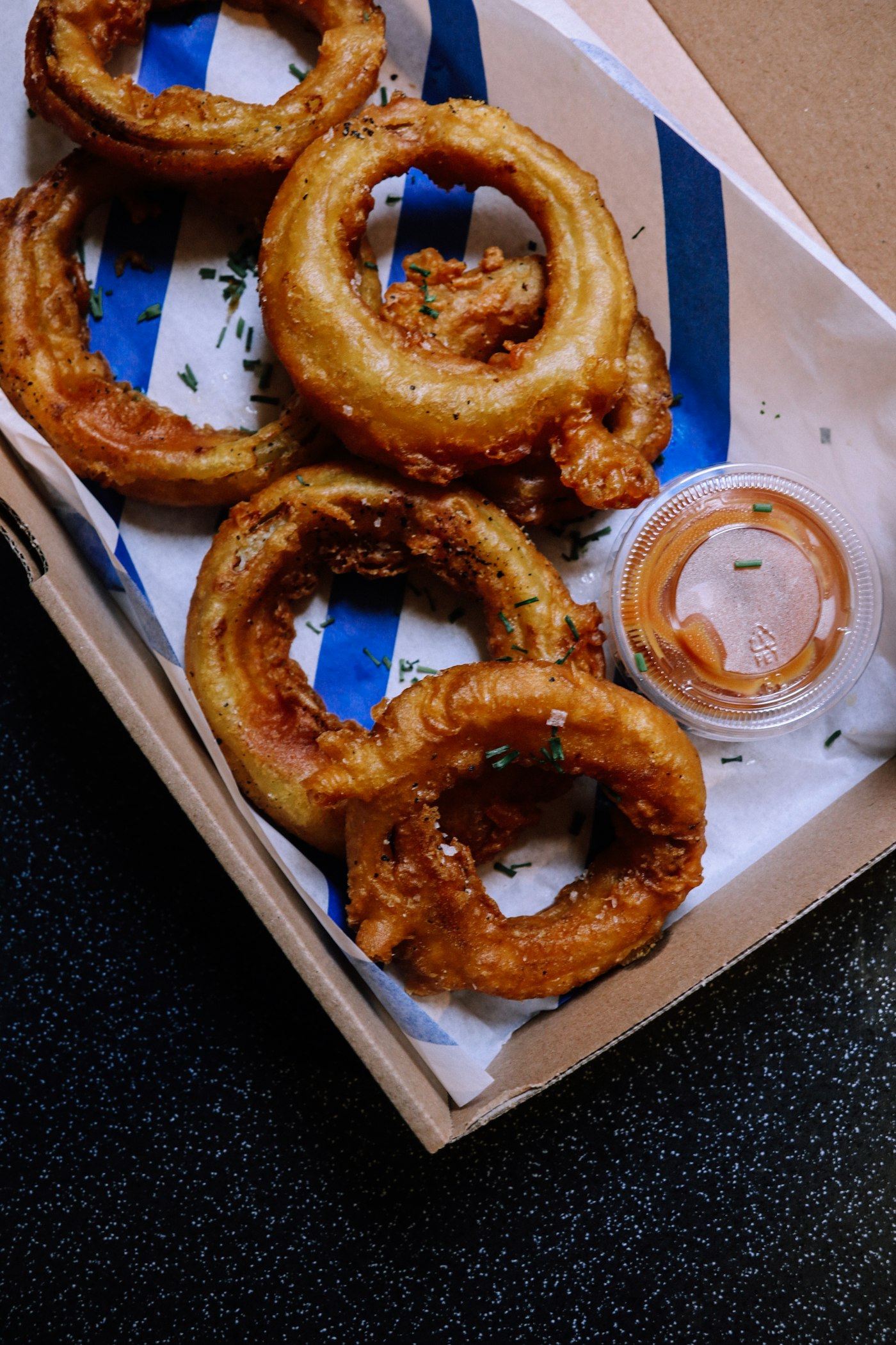 Thick battered onion rings salted and stacked in a paper lined cardboard tray with a tub of sauce.