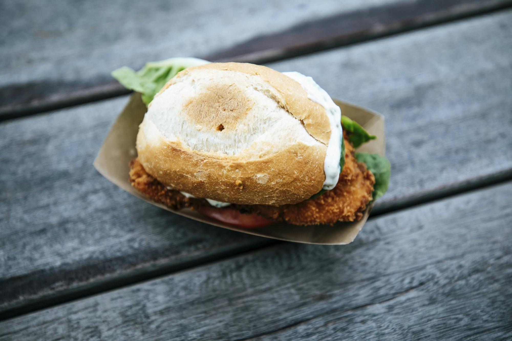 A breaded pork cutlet in a paper tray, the fried edge hanging out past every side of the bun, on a weathered grey table.