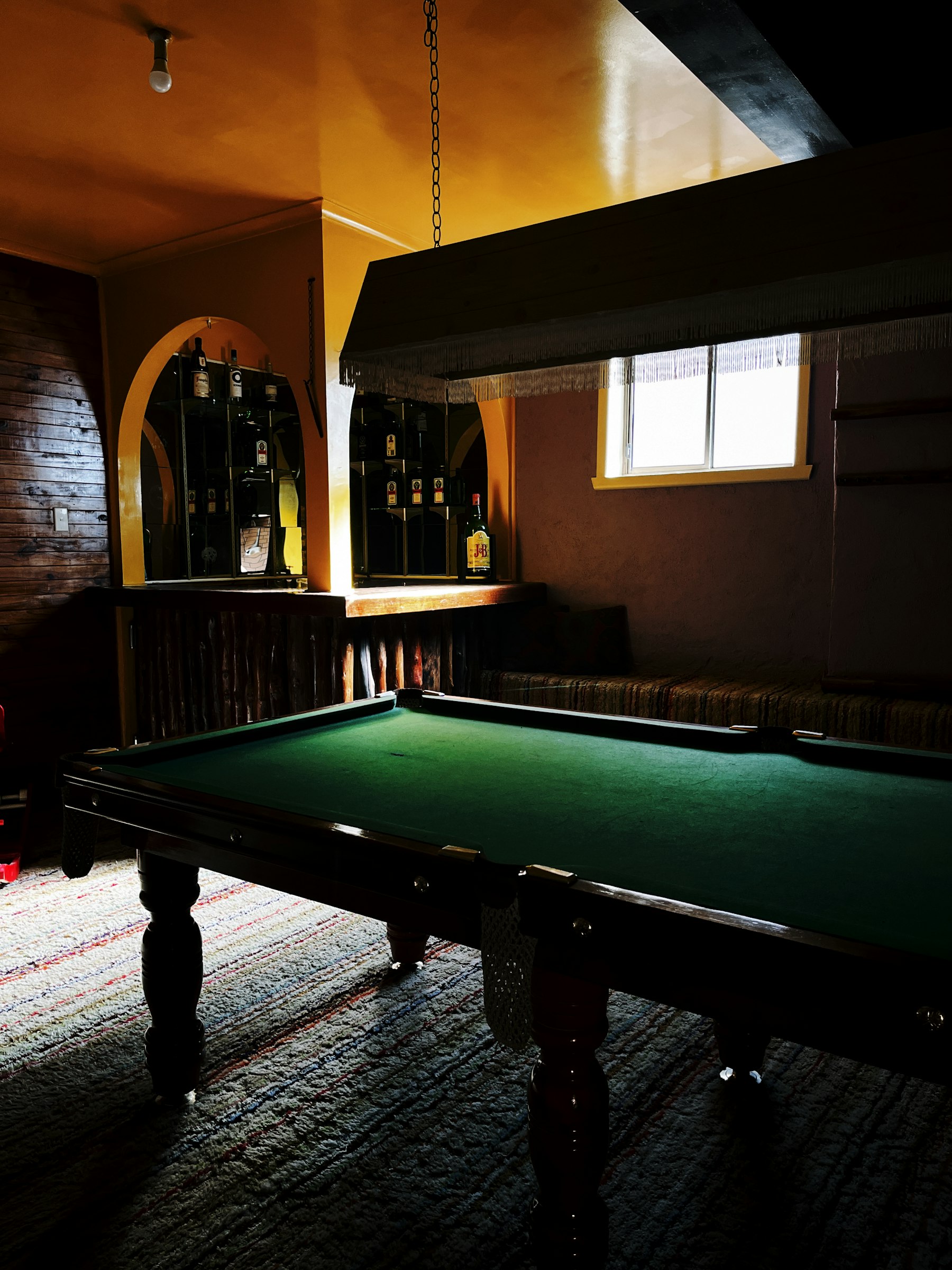 A pool table under a low hanging lamp in a dim back room, a small bar with bottles behind it and a window high on the wall.