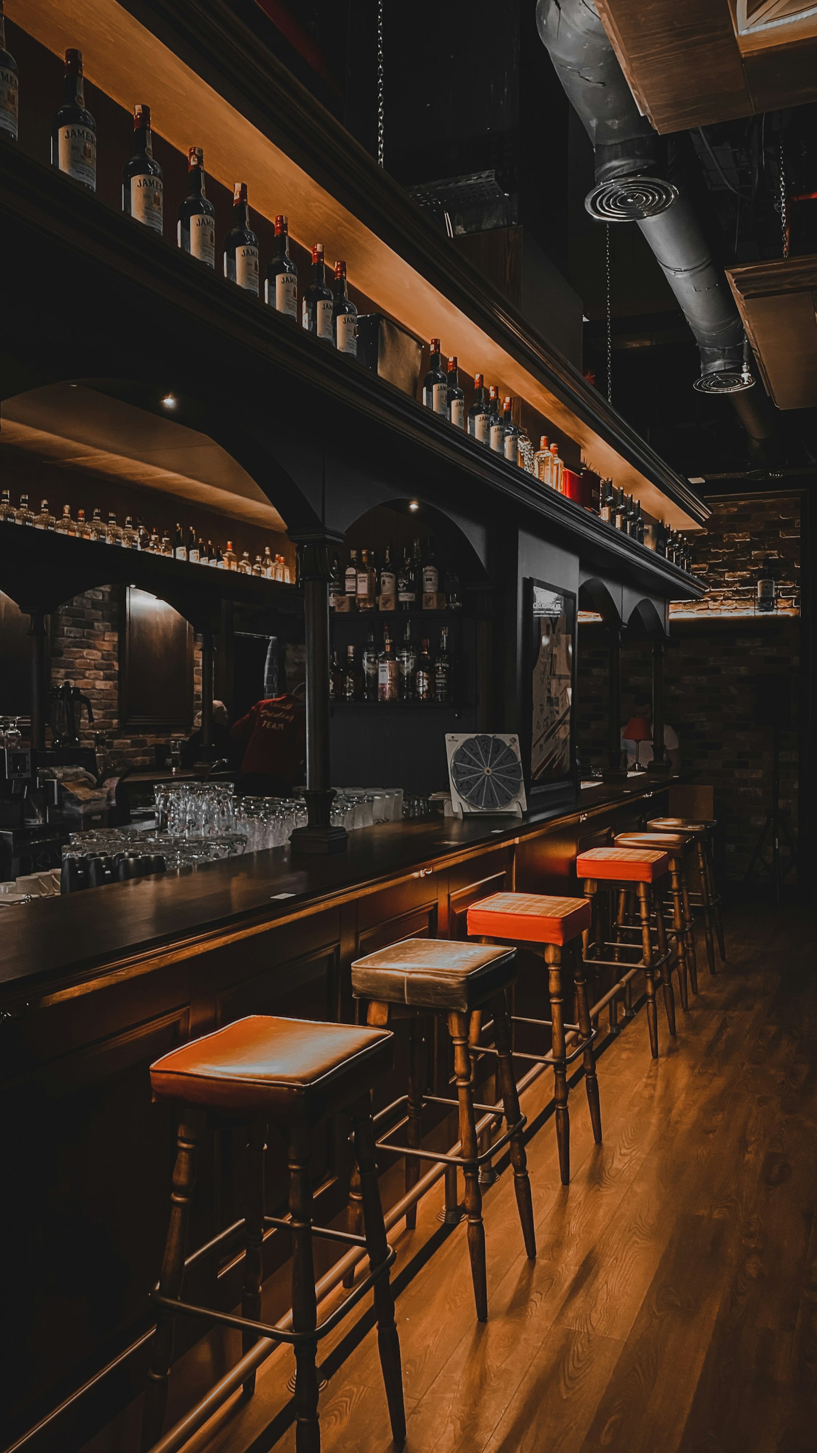 A long wooden bar in low light with orange topped stools along it and shelves of bottles lit from above.