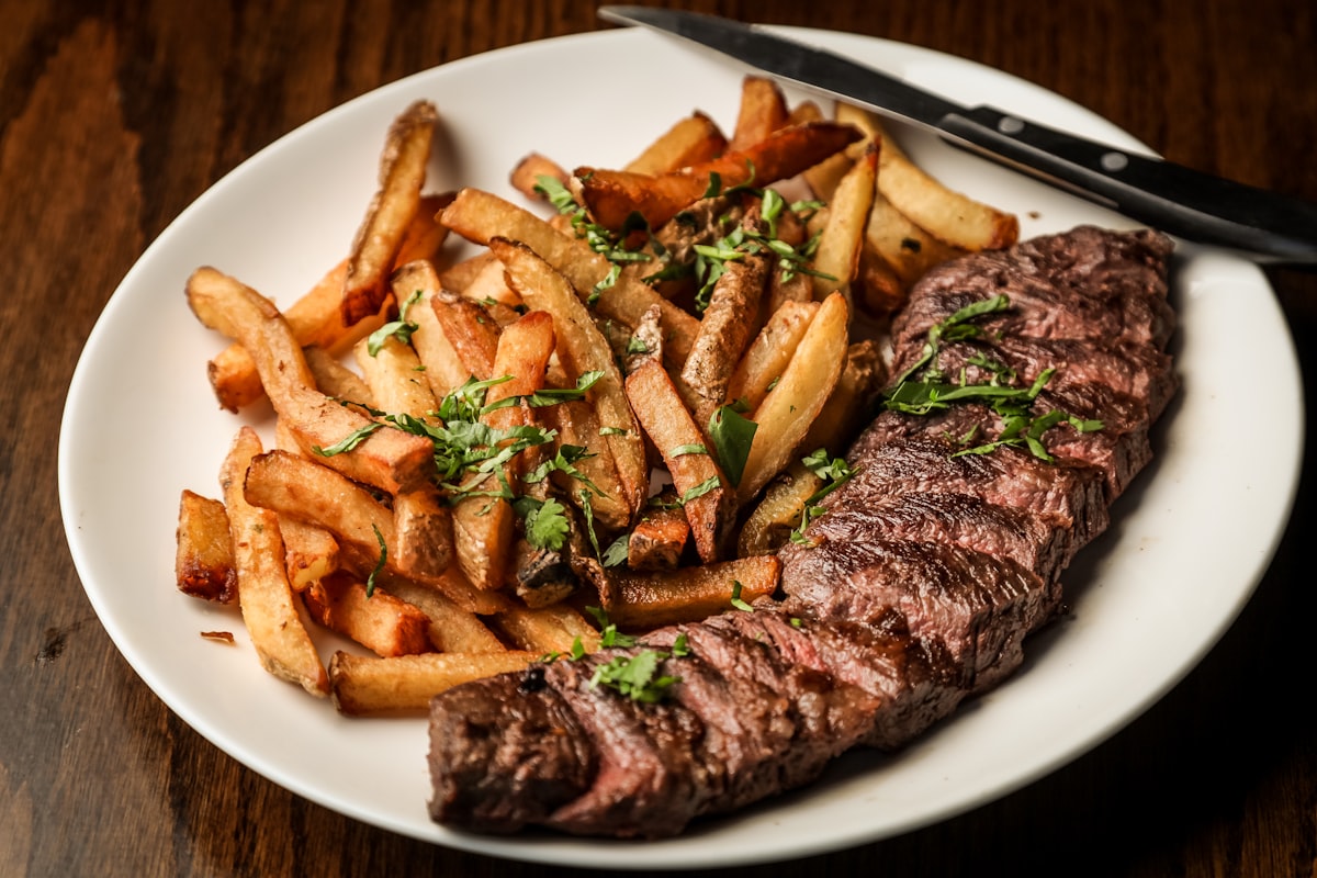 A grilled steak resting on a white plate beside a pile of fried potatoes, cut to show the middle.
