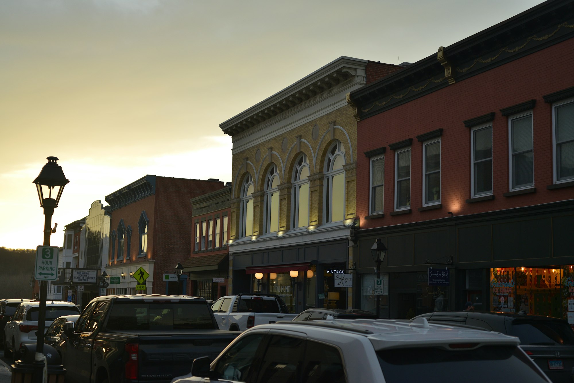 A small town main street at dusk, brick storefronts lit under a yellow sky, pickups parked at an angle along the kerb.